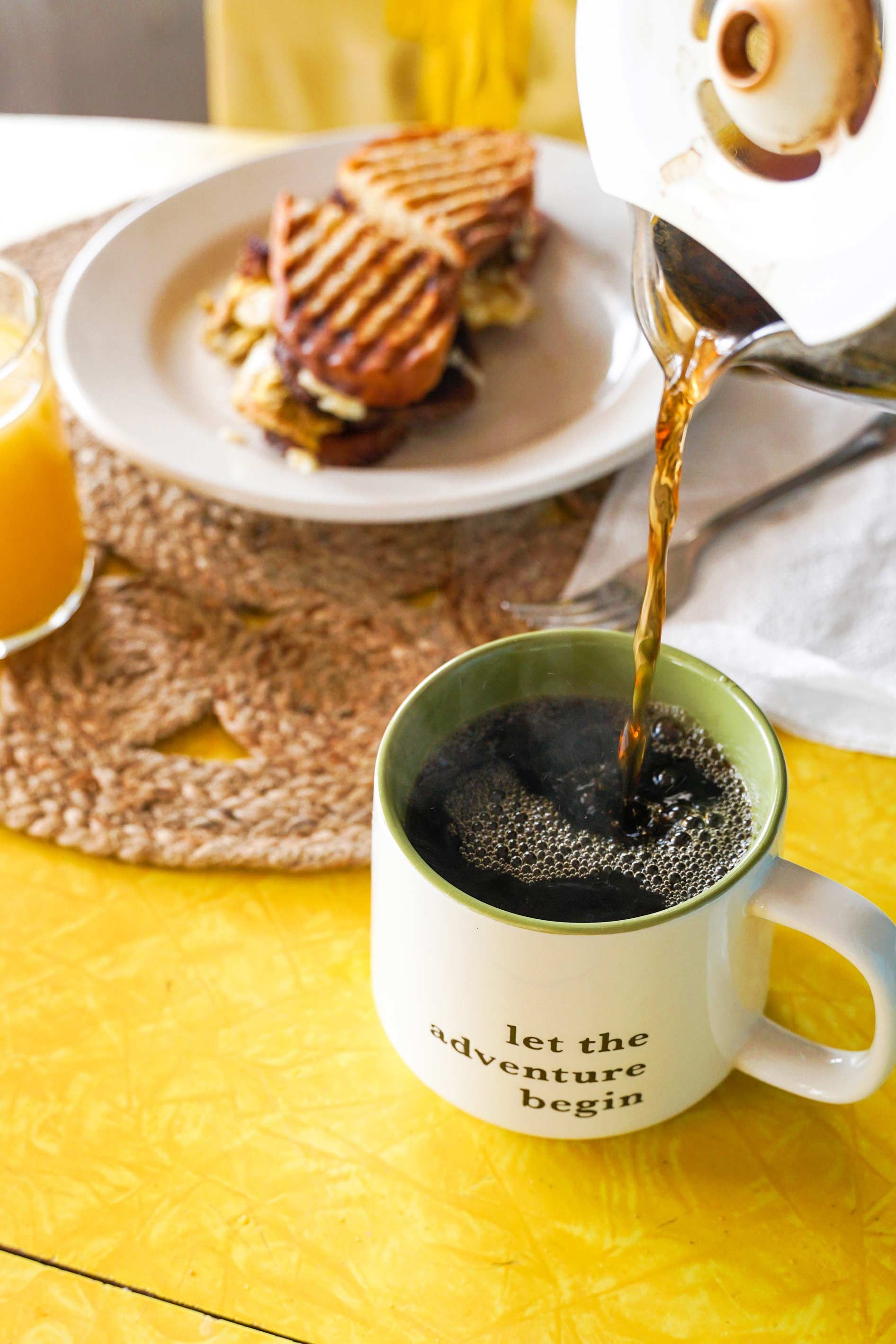 A cup of coffee is being poured into a mug with the words let the adventure begin on it.