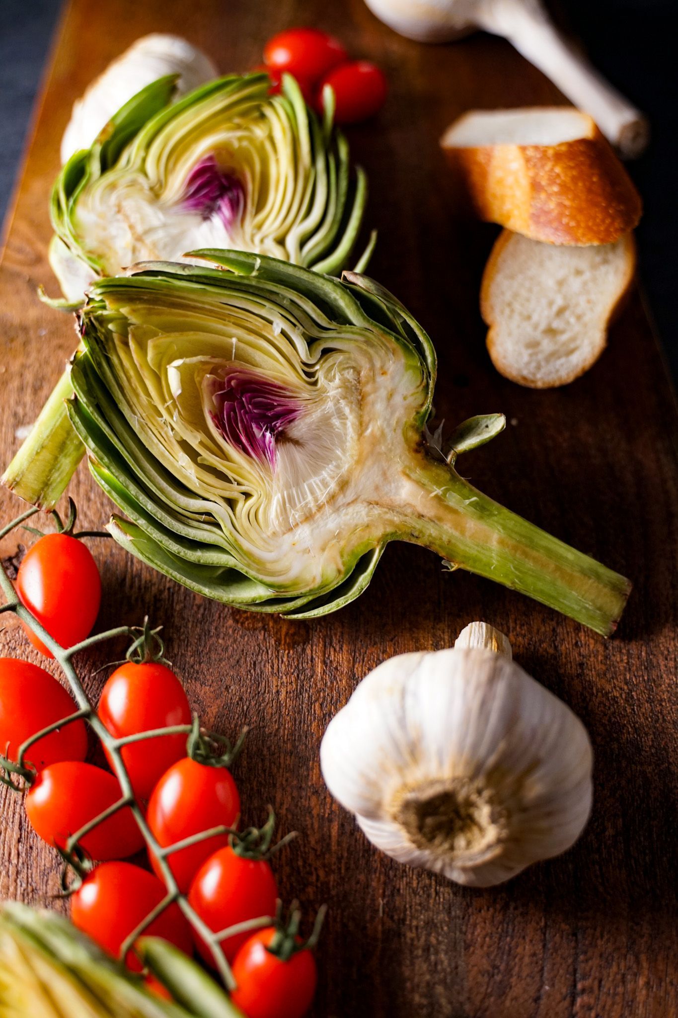 Artichokes , tomatoes , garlic and bread are on a wooden cutting board.