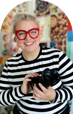 A woman wearing glasses and a striped shirt is holding a camera and smiling.