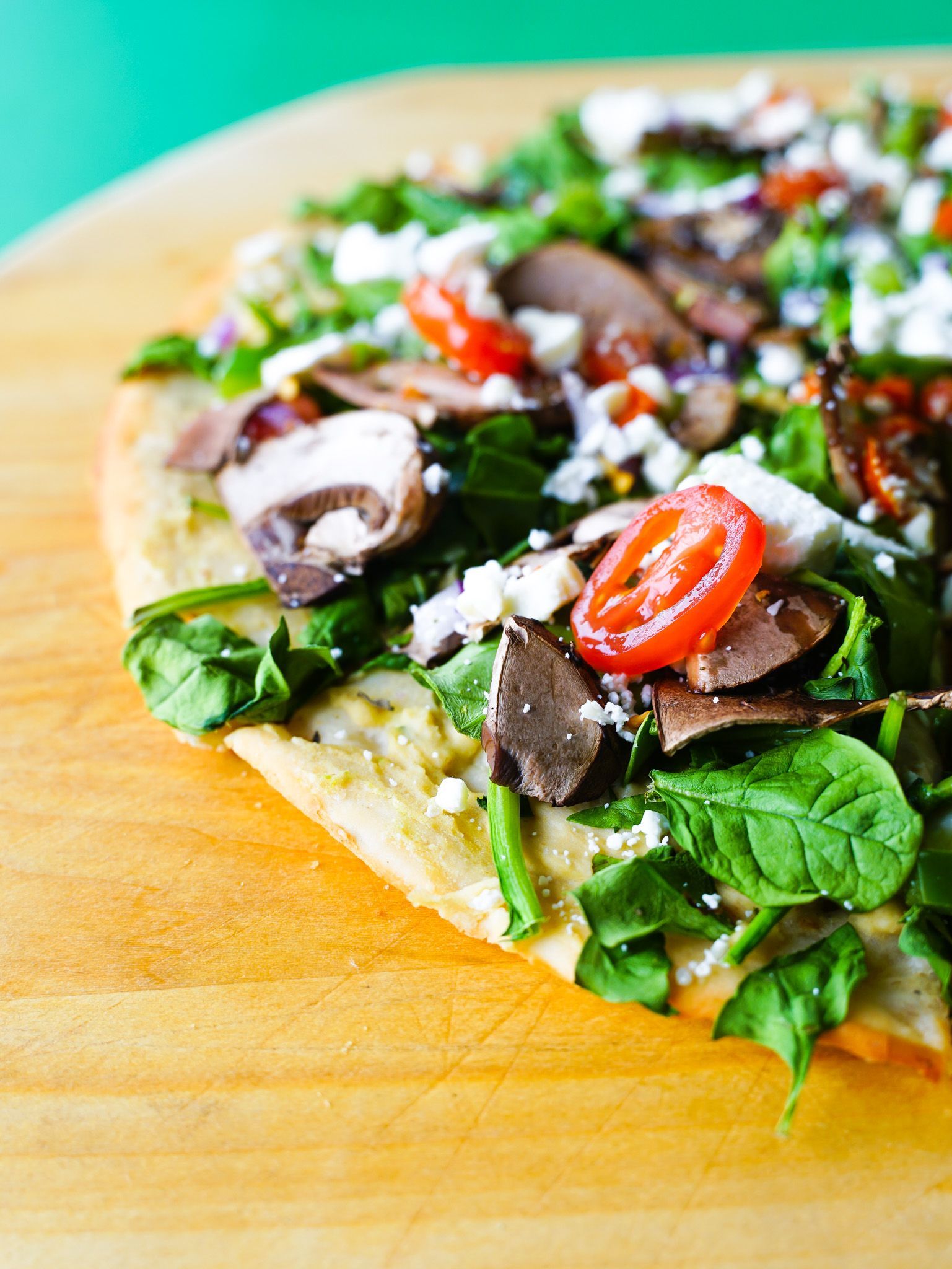 A pizza with spinach , mushrooms , tomatoes and feta cheese on a wooden cutting board.