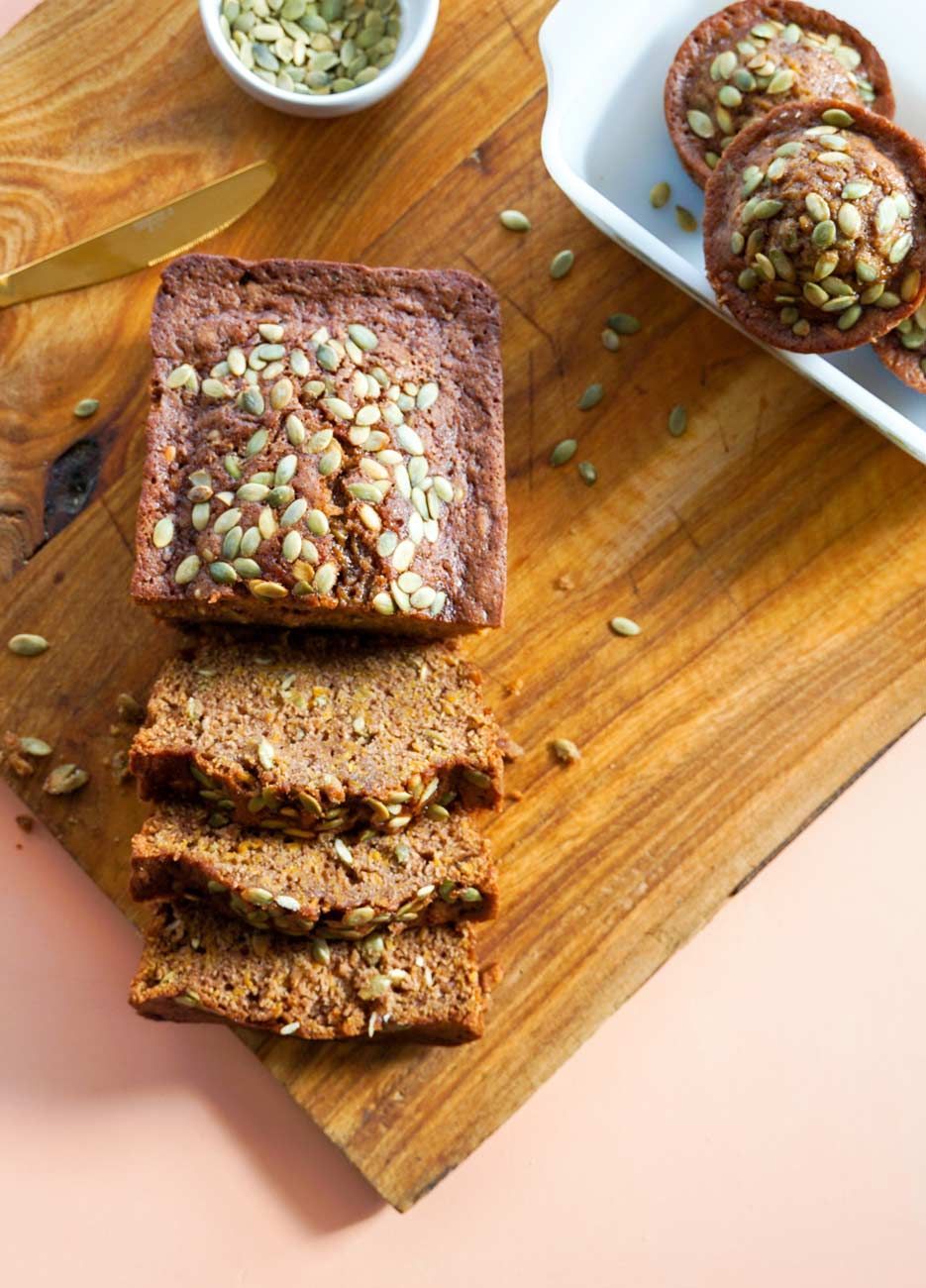 A loaf of bread is sitting on a wooden cutting board next to a bowl of pumpkin seeds.