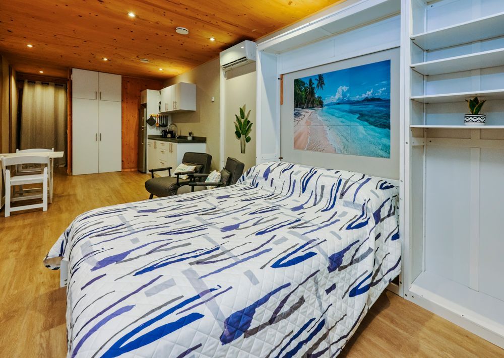 A studio apartment with a bed featuring a blue and white patterned quilt, next to built-in white shelving and a kitchen.