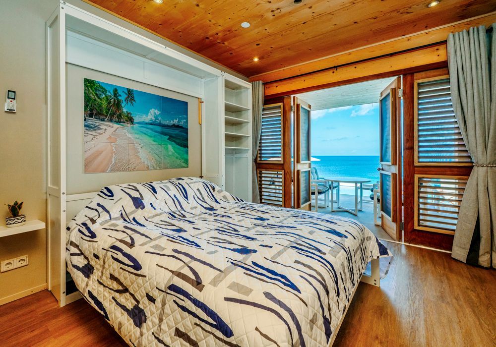 A modern bedroom with a Murphy bed, patterned quilt, and large glass doors opening to a scenic ocean view.