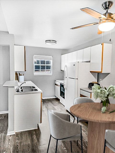 White kitchen with cabinets and appliances, small dining table, wooden floor, and ceiling fan.