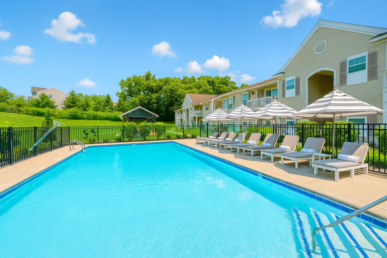Swimming pool with lounge chairs and umbrellas in front of a light-colored building under a blue sky.