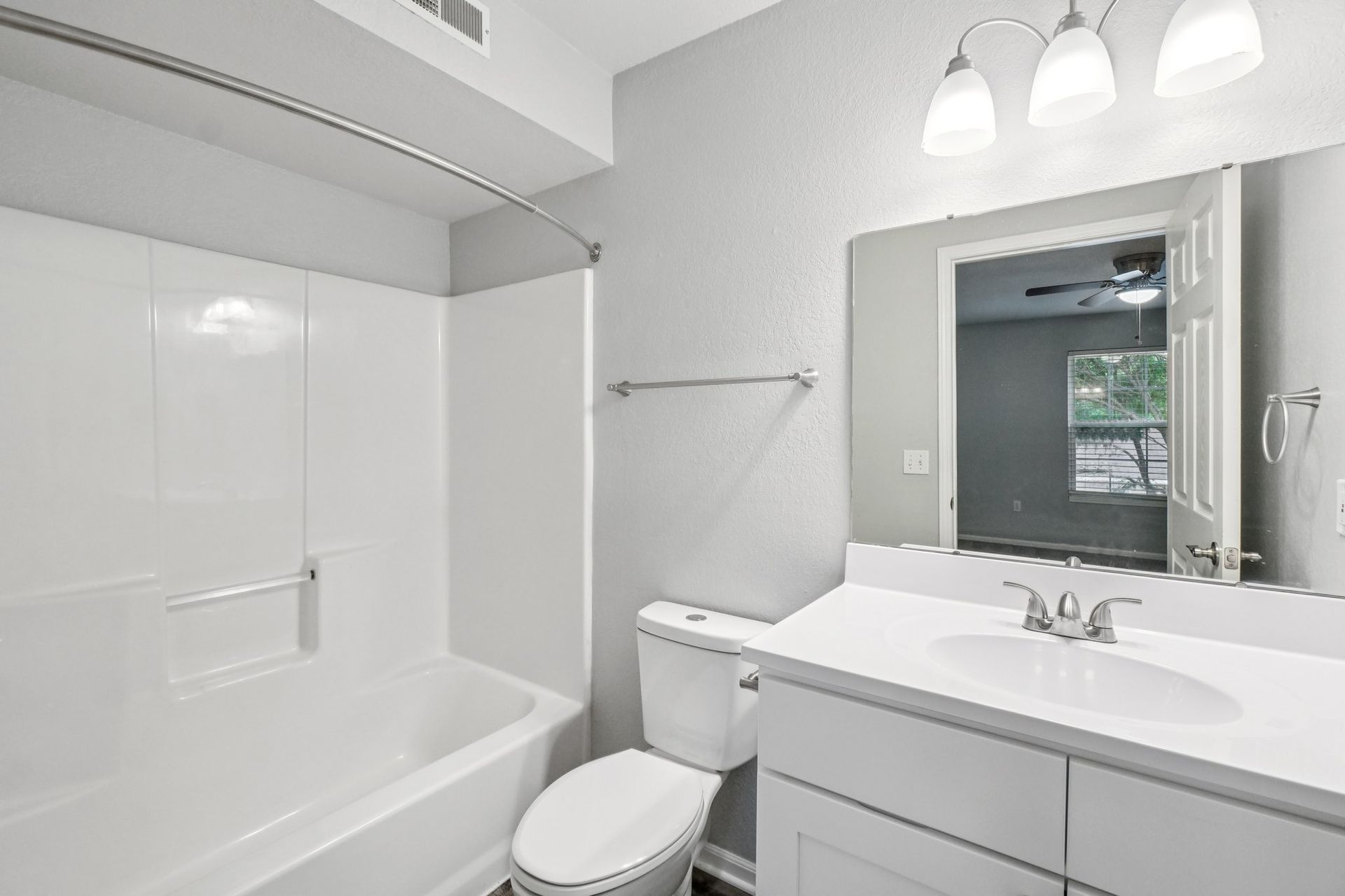 Bathroom with white tub, toilet, vanity, and gray walls. A mirror reflects a bedroom and open door.