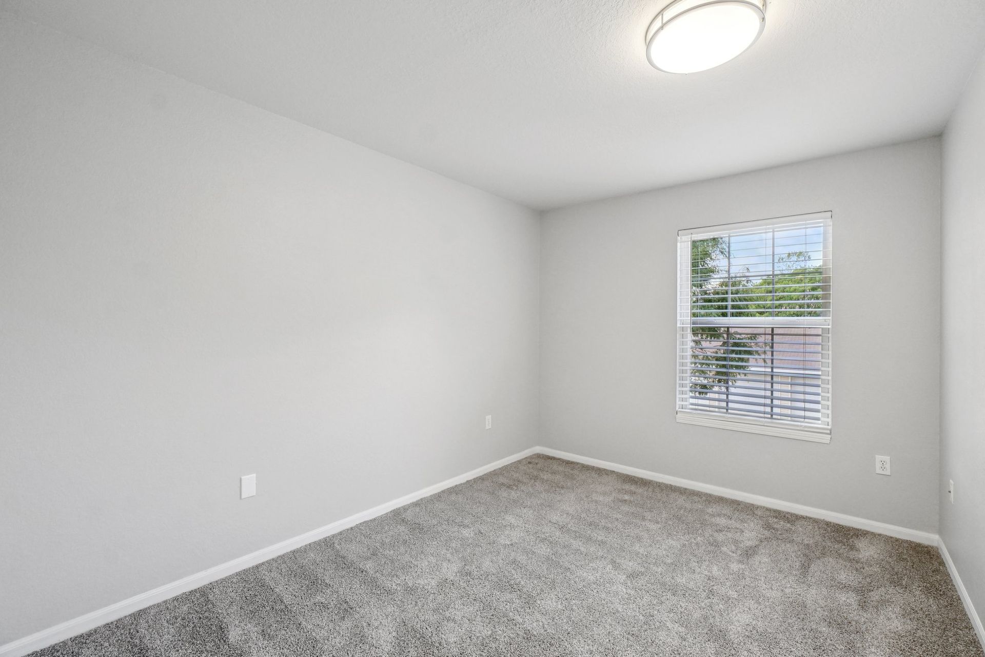 Empty, light gray bedroom with gray carpet, a small window, and a ceiling light.