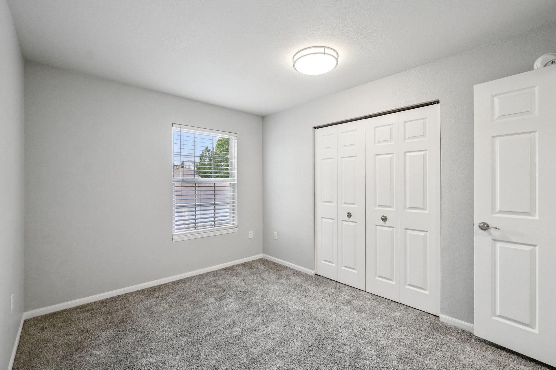 Empty bedroom with gray walls, carpet, and white closet doors. A small window lets in light.
