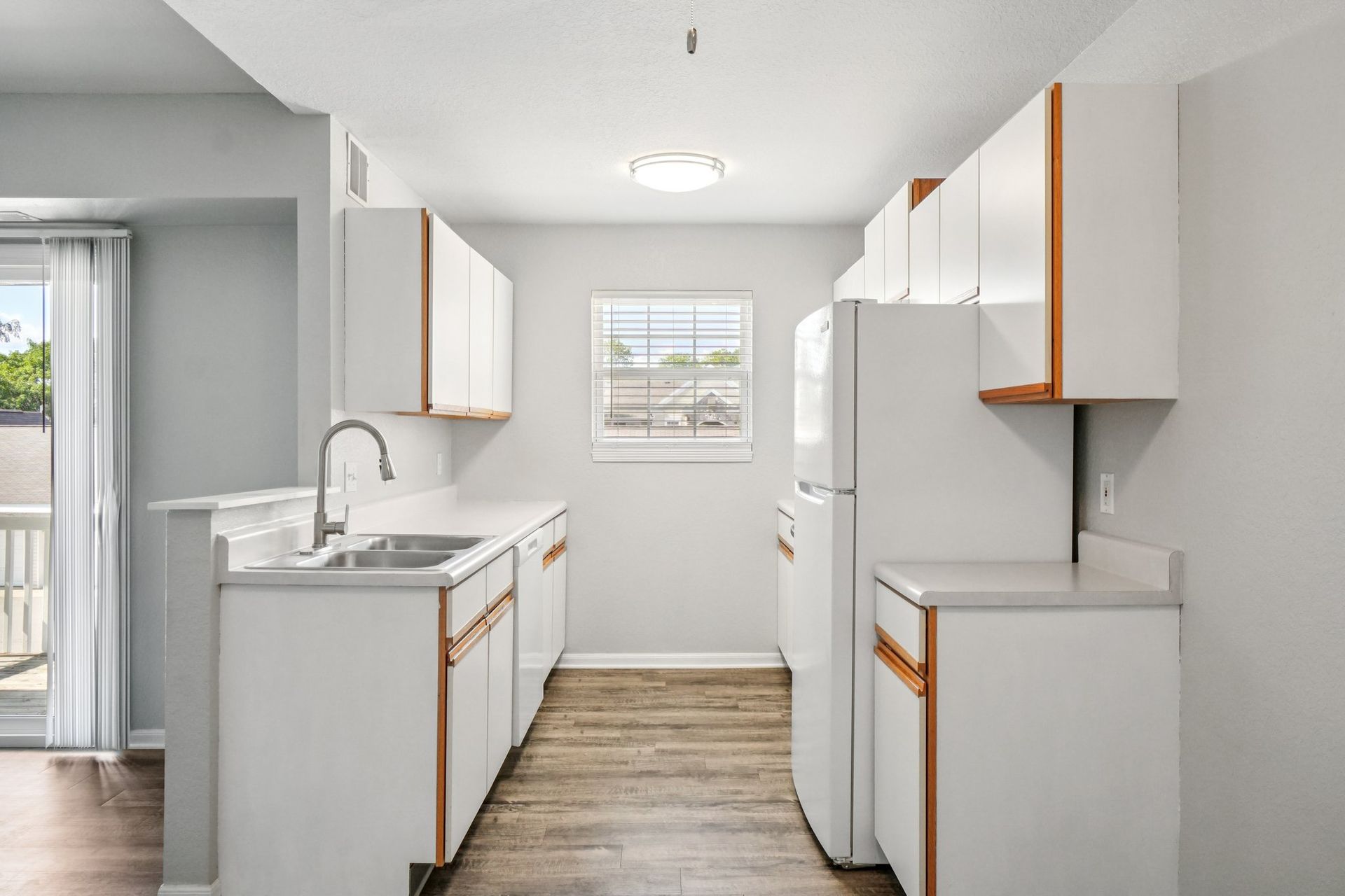 A galley kitchen with white cabinets, countertops, and appliances. A window and a sliding door are visible.