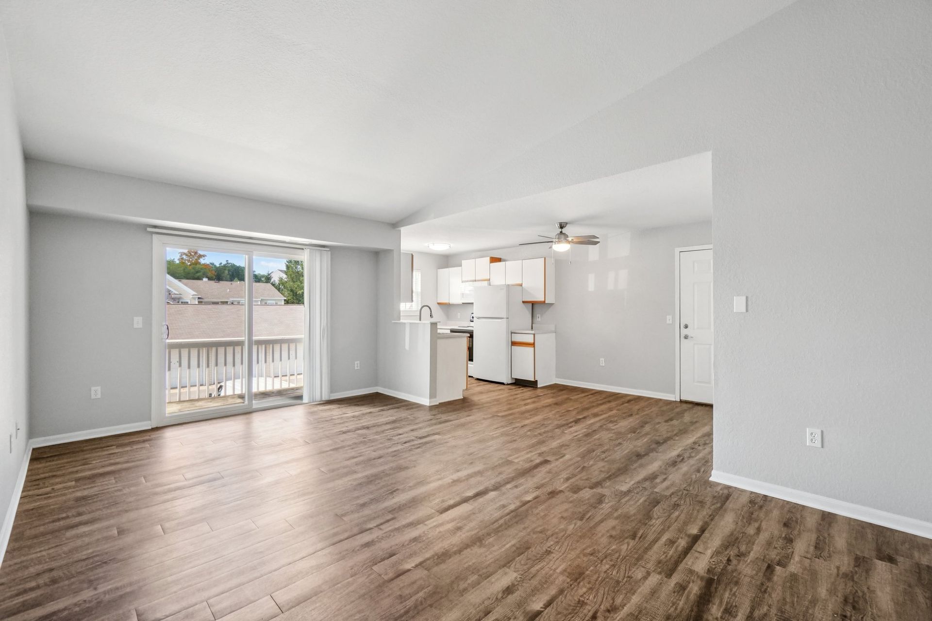Empty living room with hardwood floors, sliding glass door to balcony, and a glimpse of the kitchen.