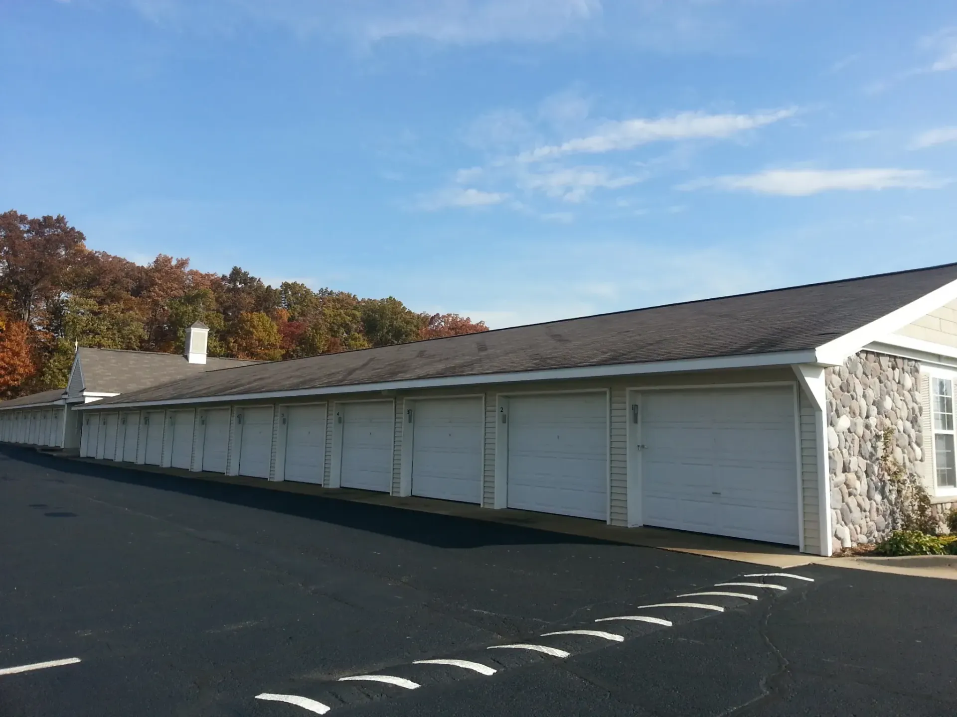 Row of white garages with dark roof, asphalt driveway, and trees with autumn leaves against a blue sky.