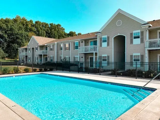 Apartment complex with a blue swimming pool and surrounding black fence, under a clear blue sky.