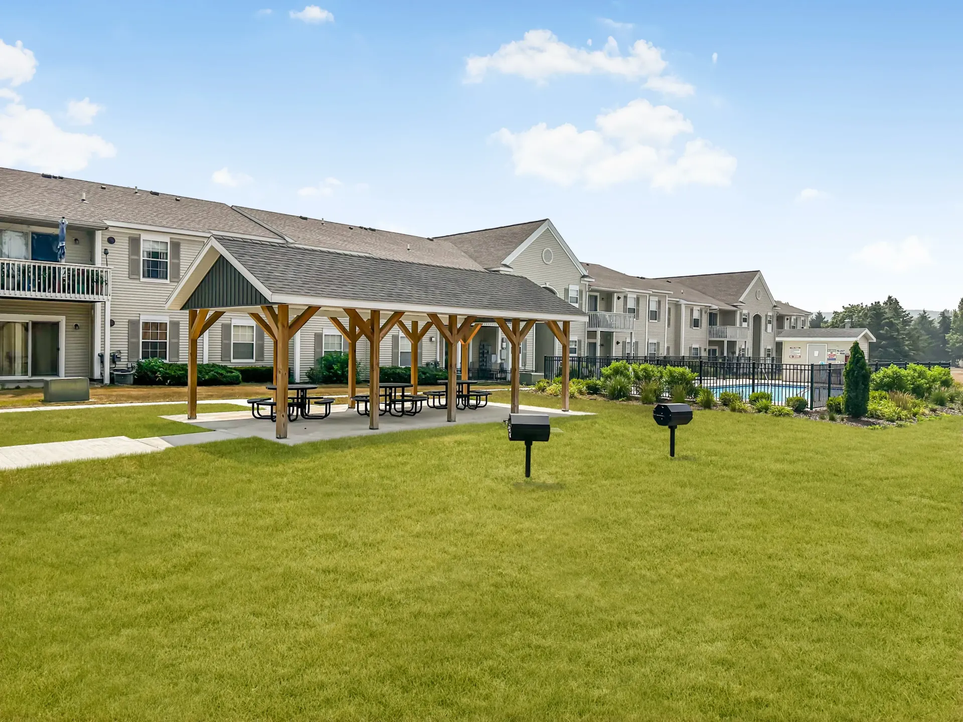 Lush green lawn with picnic shelter, grills, and apartments in the background near a pool. Sunny day.