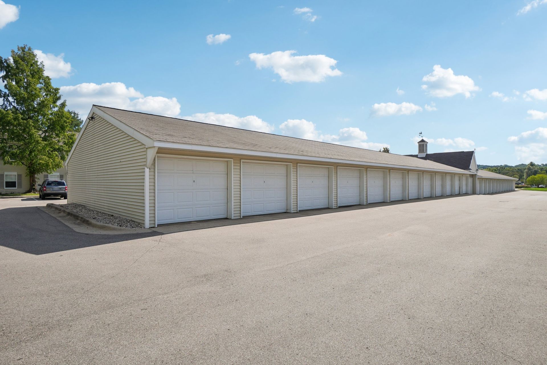 Row of storage units with white doors under a beige building, on a gray asphalt lot, under blue sky with clouds.