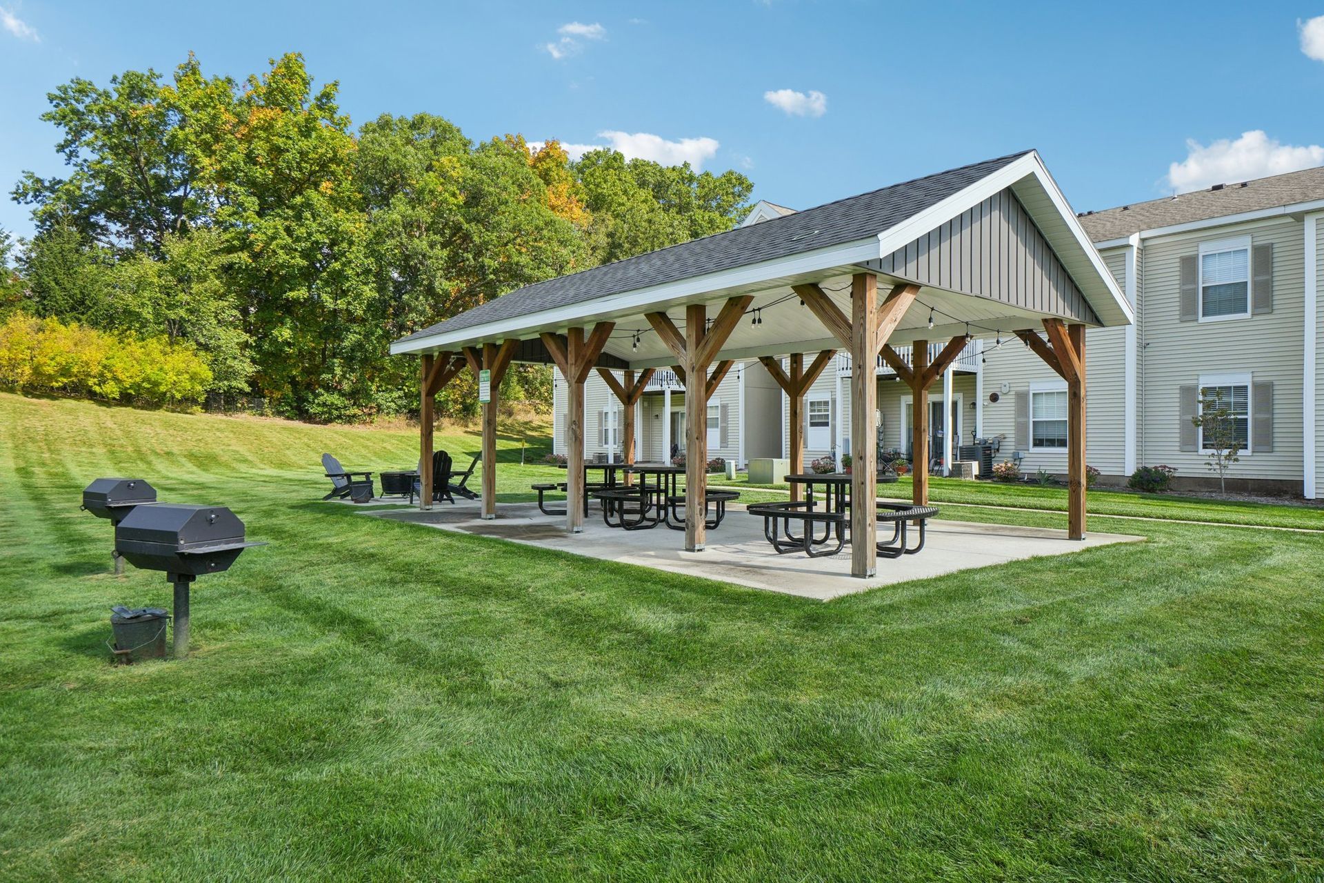 Covered picnic area with tables, grills, and green lawn. Buildings and trees in the background.