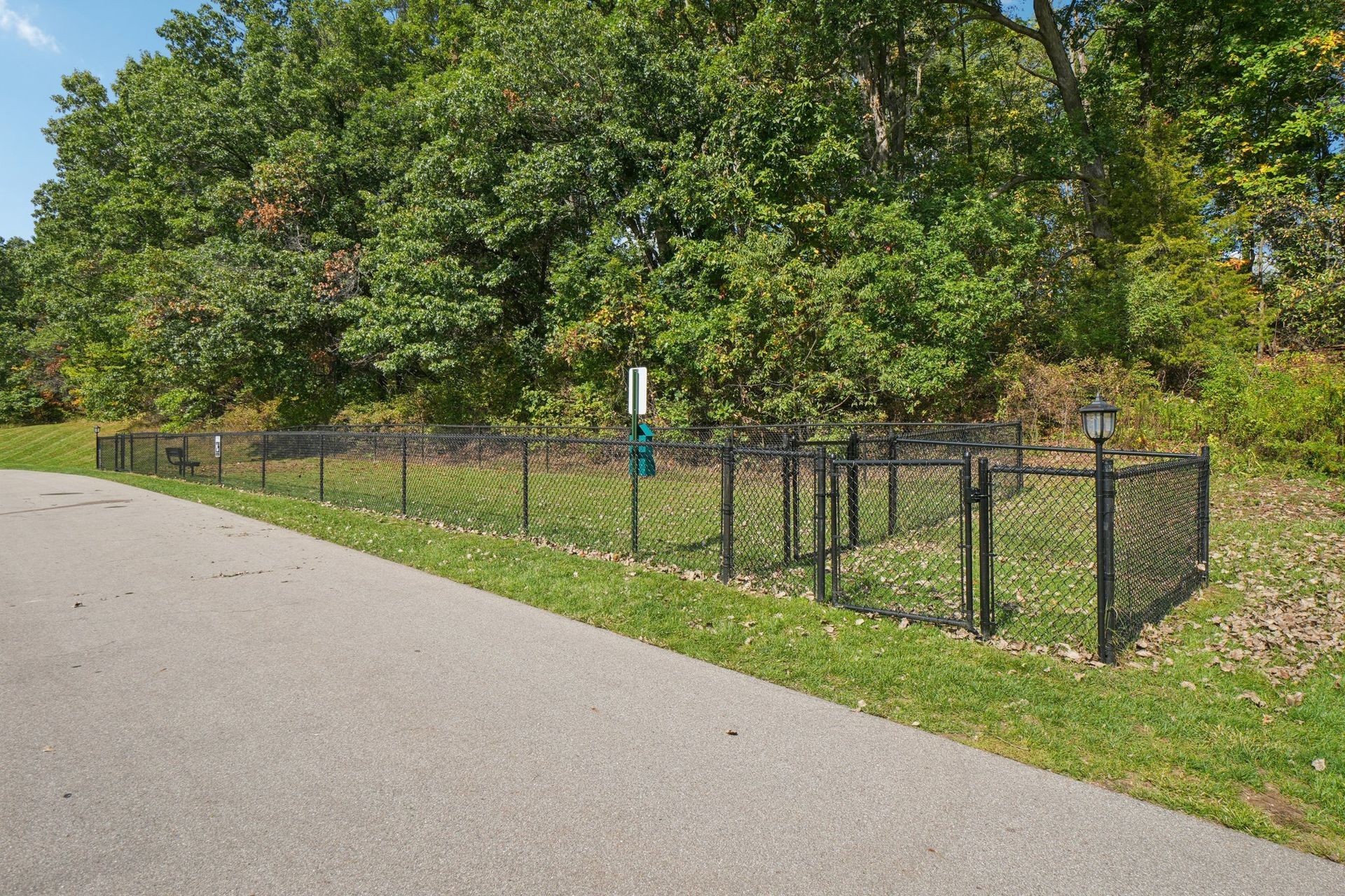Black chain-link fence with dog waste station next to path, trees in the background.