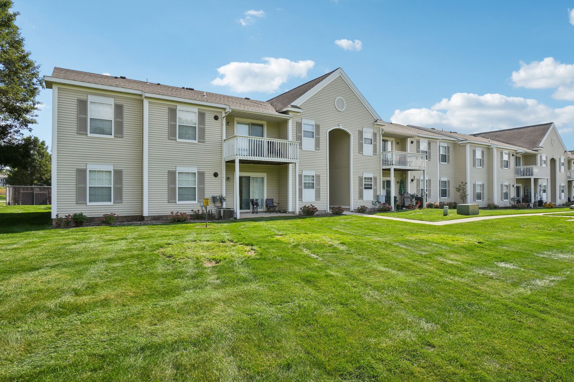 Apartment building exterior, light-colored siding, multiple units with balconies, green lawn, sunny day.
