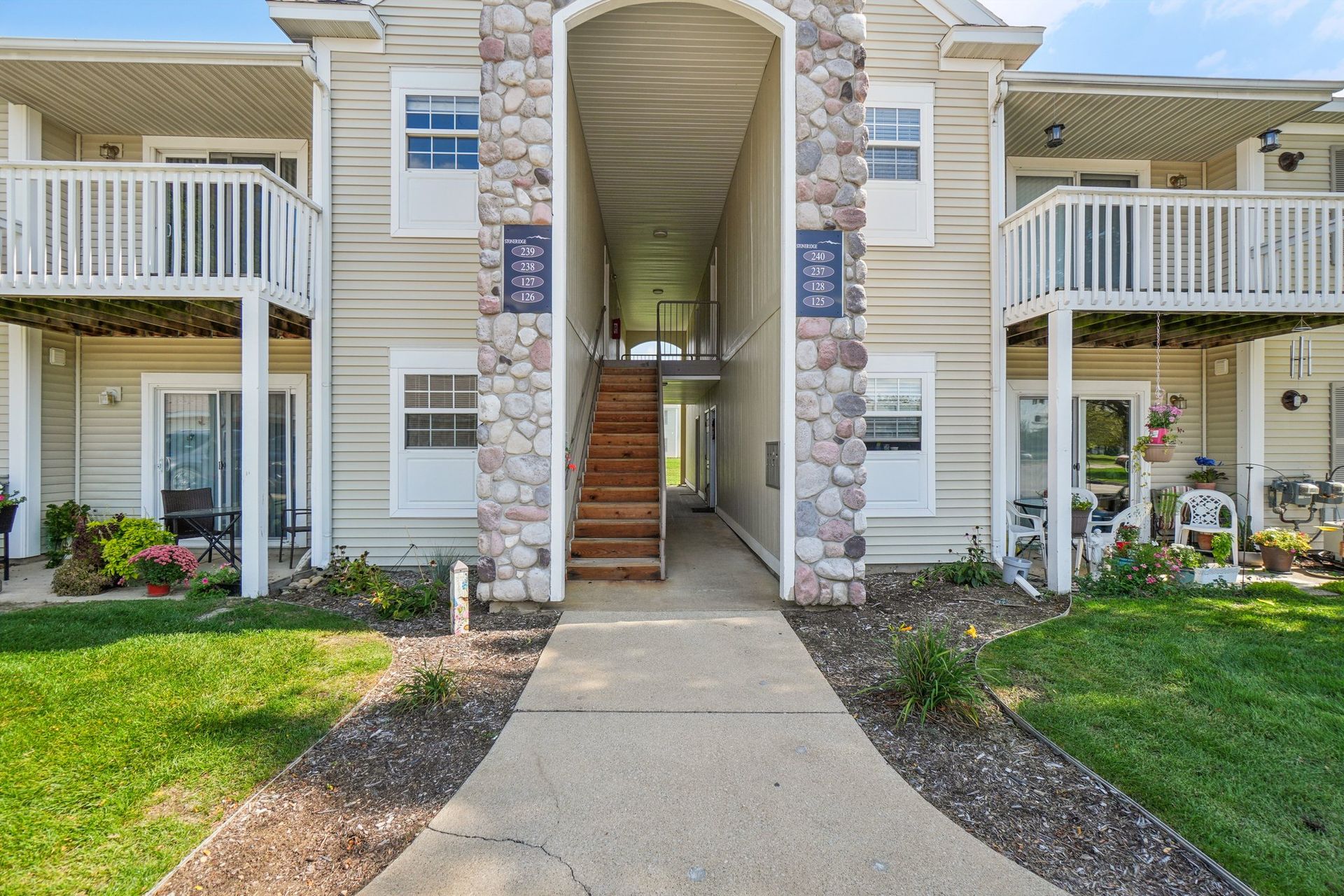 Exterior view of two-story tan apartment building. Stone archway leads to stairs. Balconies on the upper level, grass lawn.