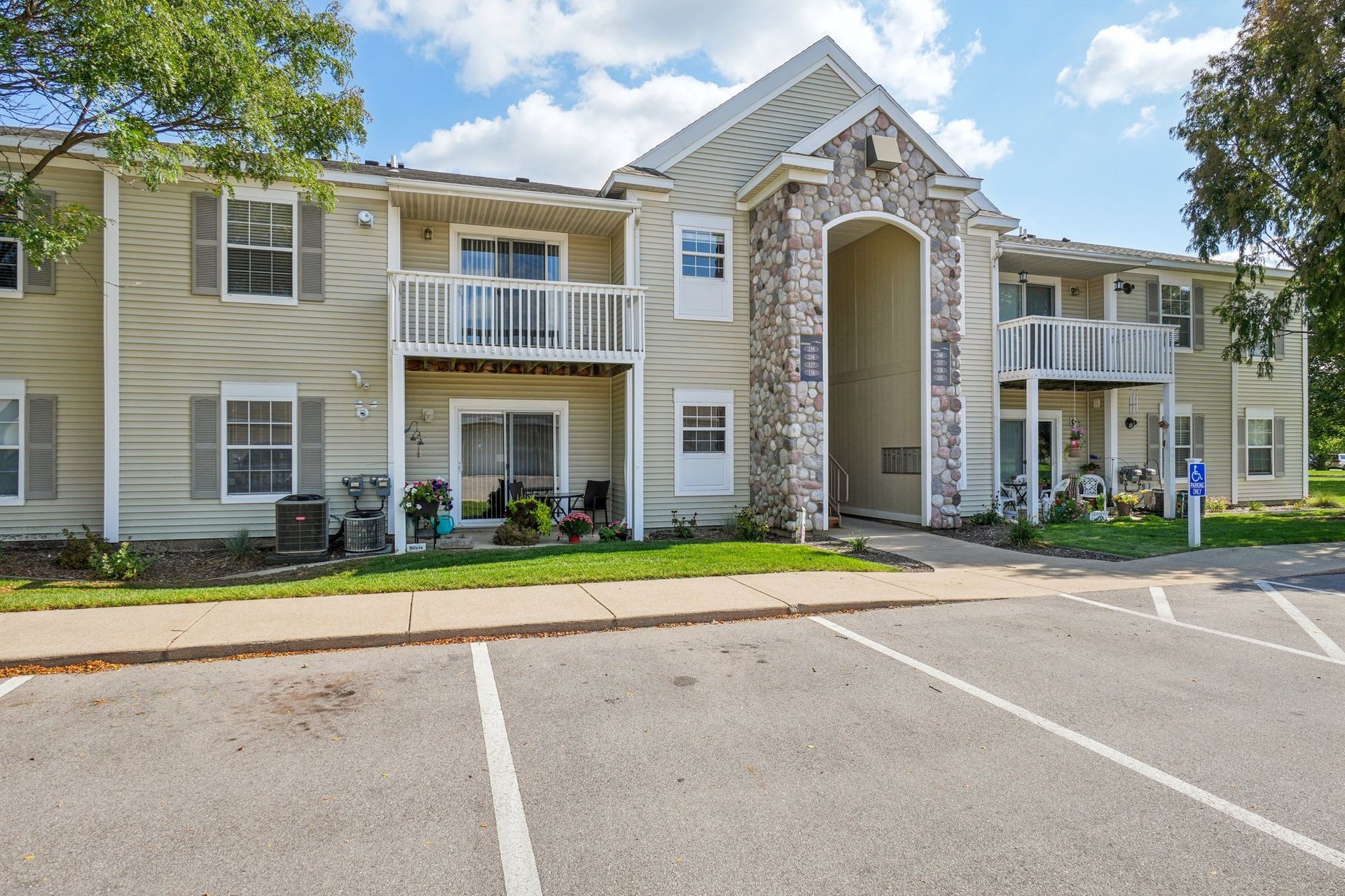 Apartment building exterior, two stories, beige siding, stone entrance, balconies, parking spaces, sunny day.