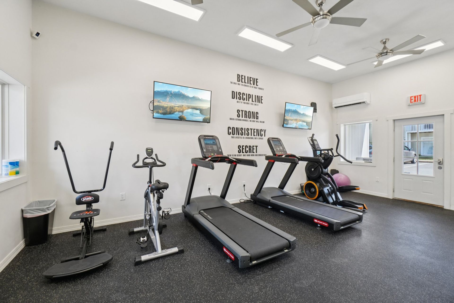 Gym interior with exercise equipment: treadmills, bikes, TVs, and inspirational text on wall. Black floor.