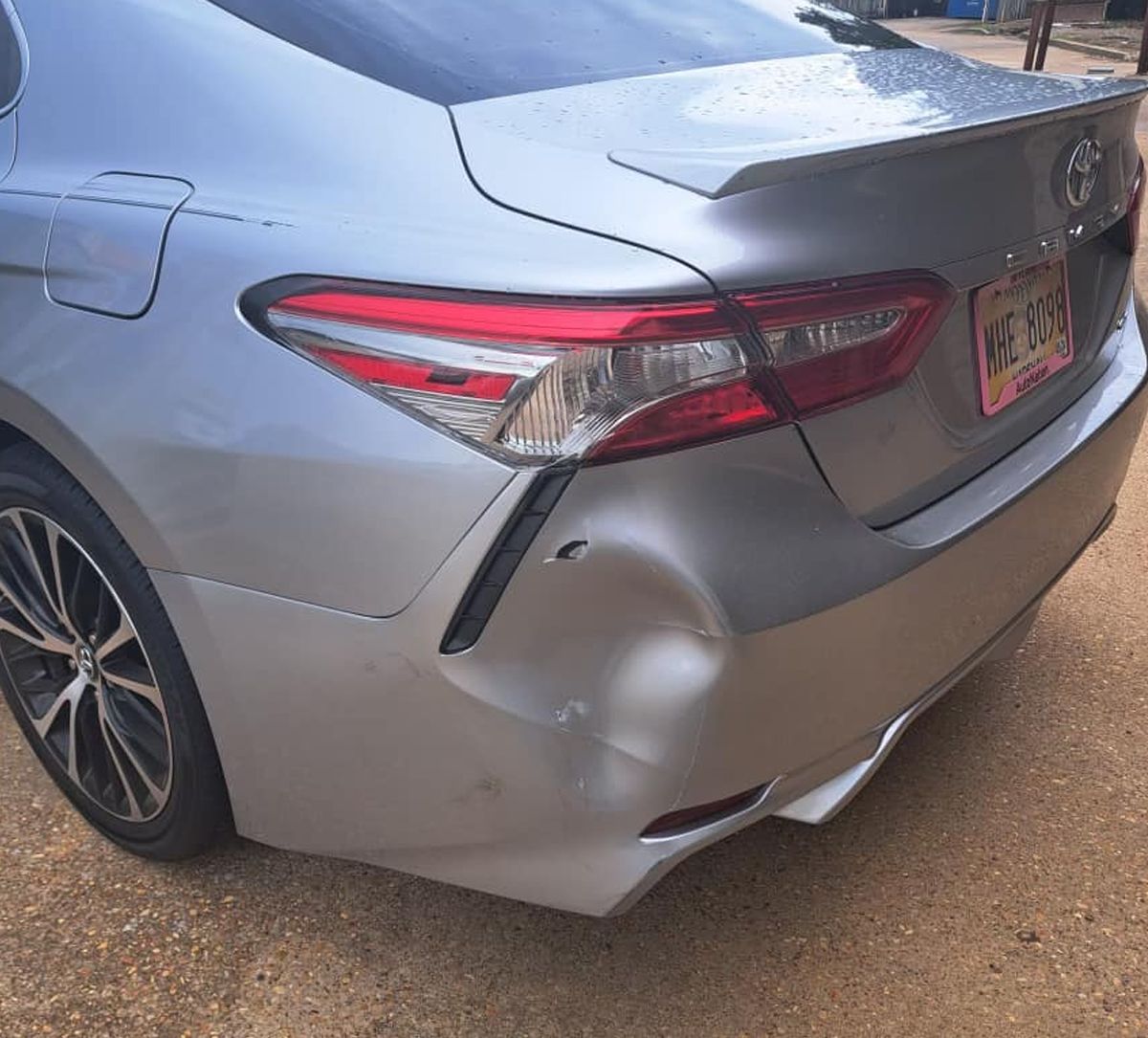 Rear side of a silver sedan with a dented bumper and damaged taillight parked on gravel.