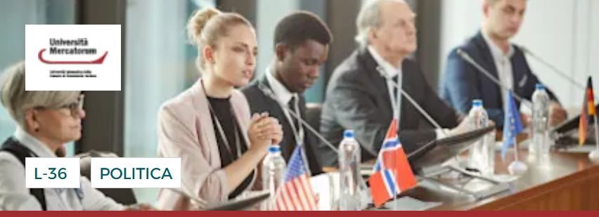 Persone sedute a un tavolo da conferenza con bandiere. Una persona sta parlando. È visibile il logo dell'università.