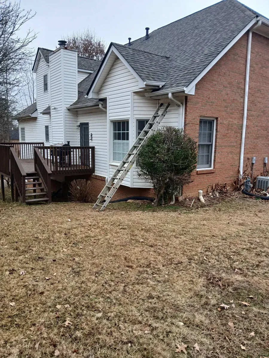 A ladder leans against a two-story house with a damaged roof and a brick exterior. A wooden deck is on the left.