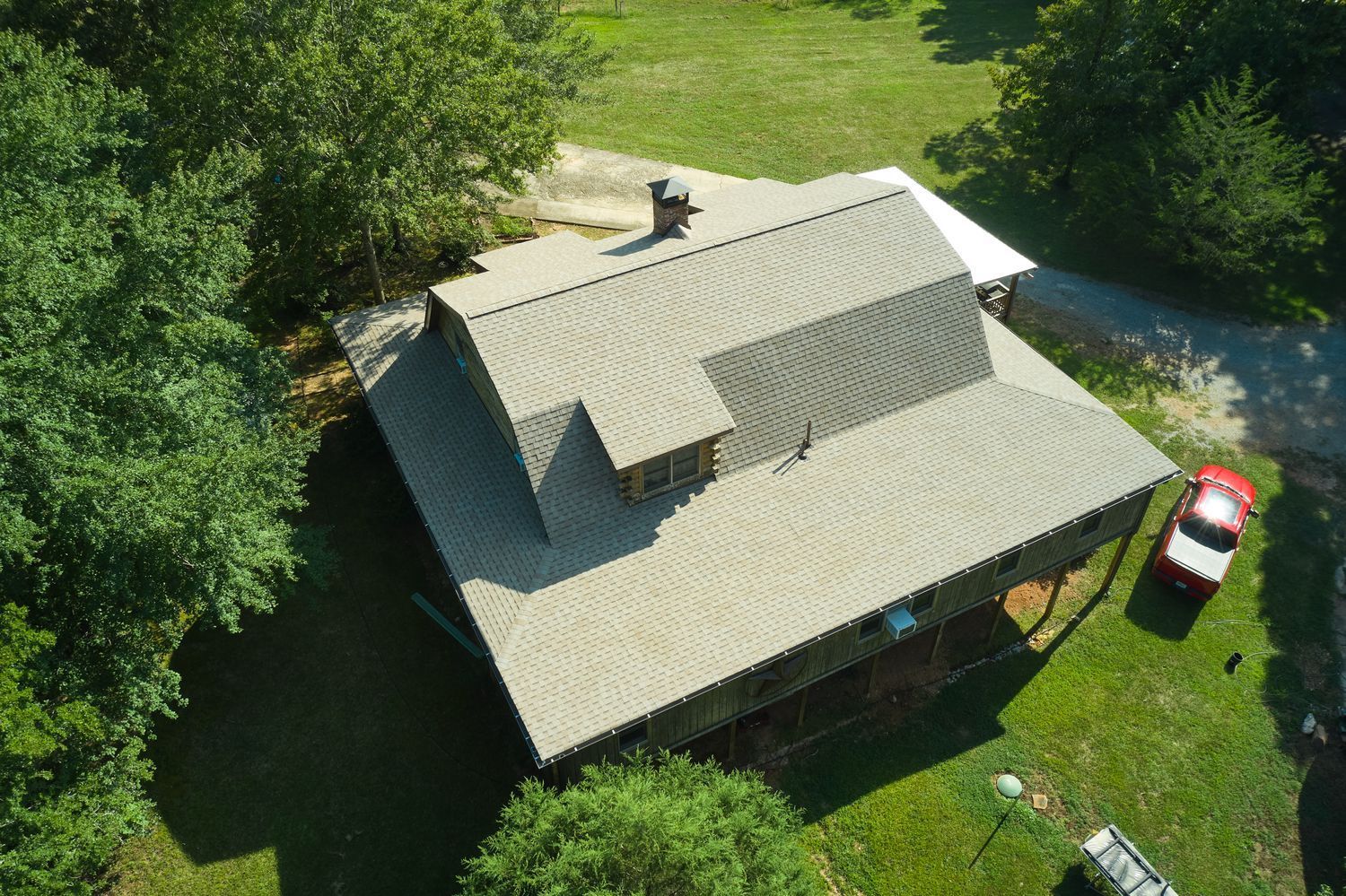 Aerial view of a house with a gray roof, surrounded by green trees and grass, with a red car parked nearby.