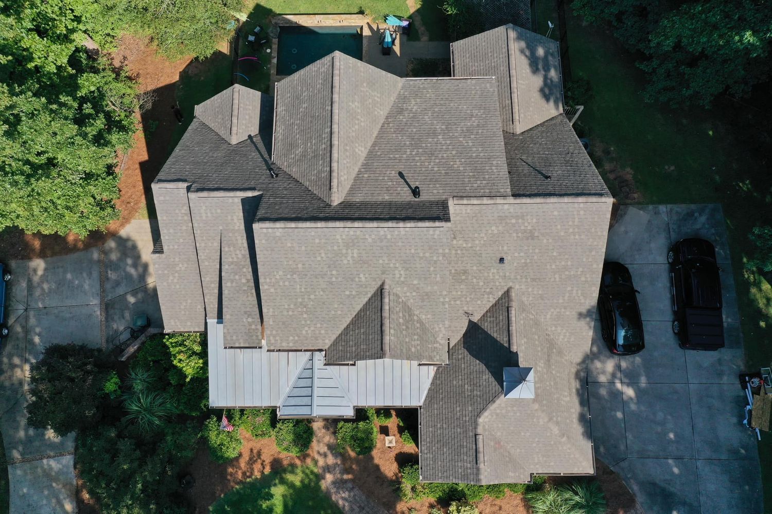 Aerial view of a gray-roofed house with a complex roofline, surrounded by trees and a driveway with cars.