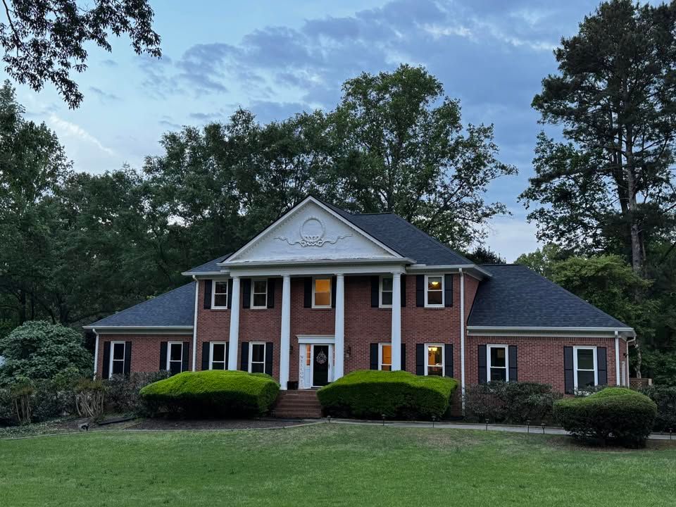 Brick two-story house with columns and a black roof, set against a backdrop of trees, with a grassy lawn.