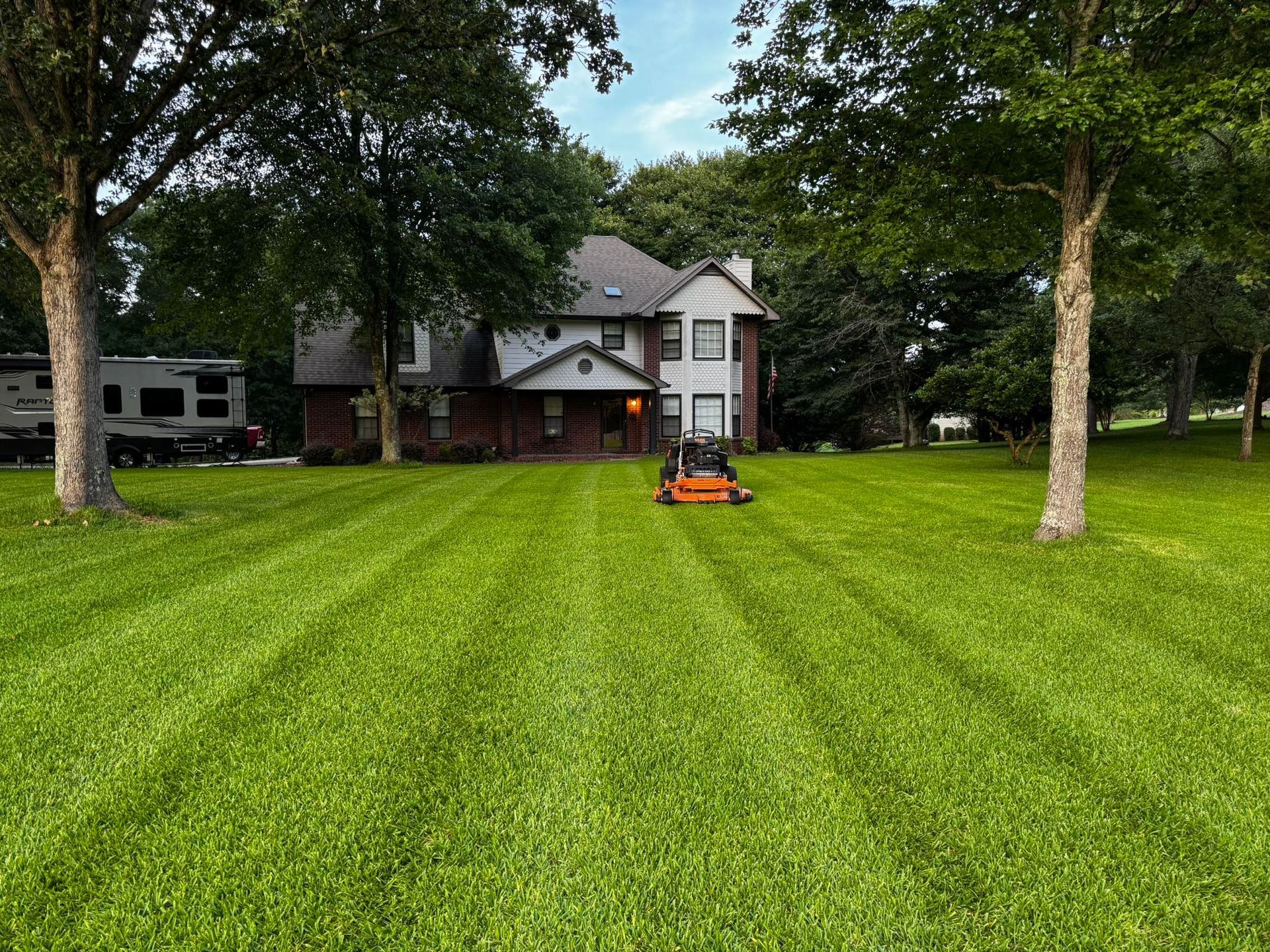 Lawn mower cutting stripes in a large green lawn in front of a two-story house with trees.