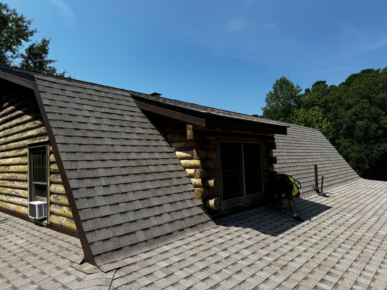 Log cabin roof with asphalt shingles; person in yellow vest on roof under blue sky.