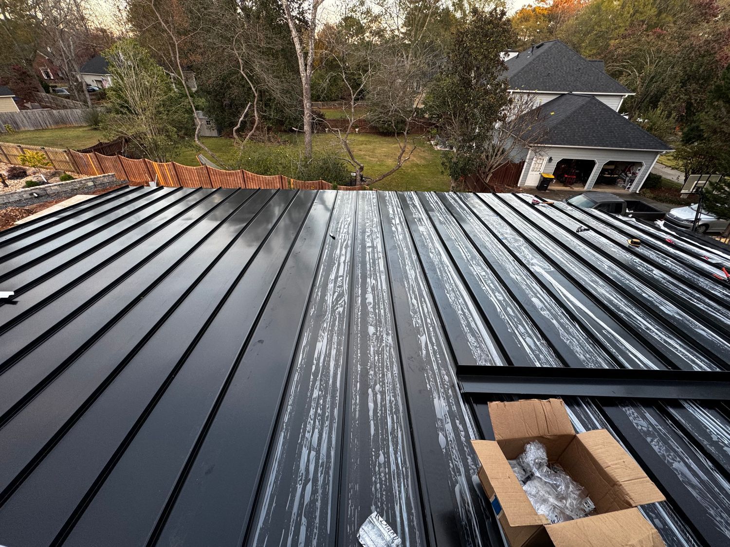 Black metal roof with exposed fasteners, partially installed, box with parts. Background with trees, houses.