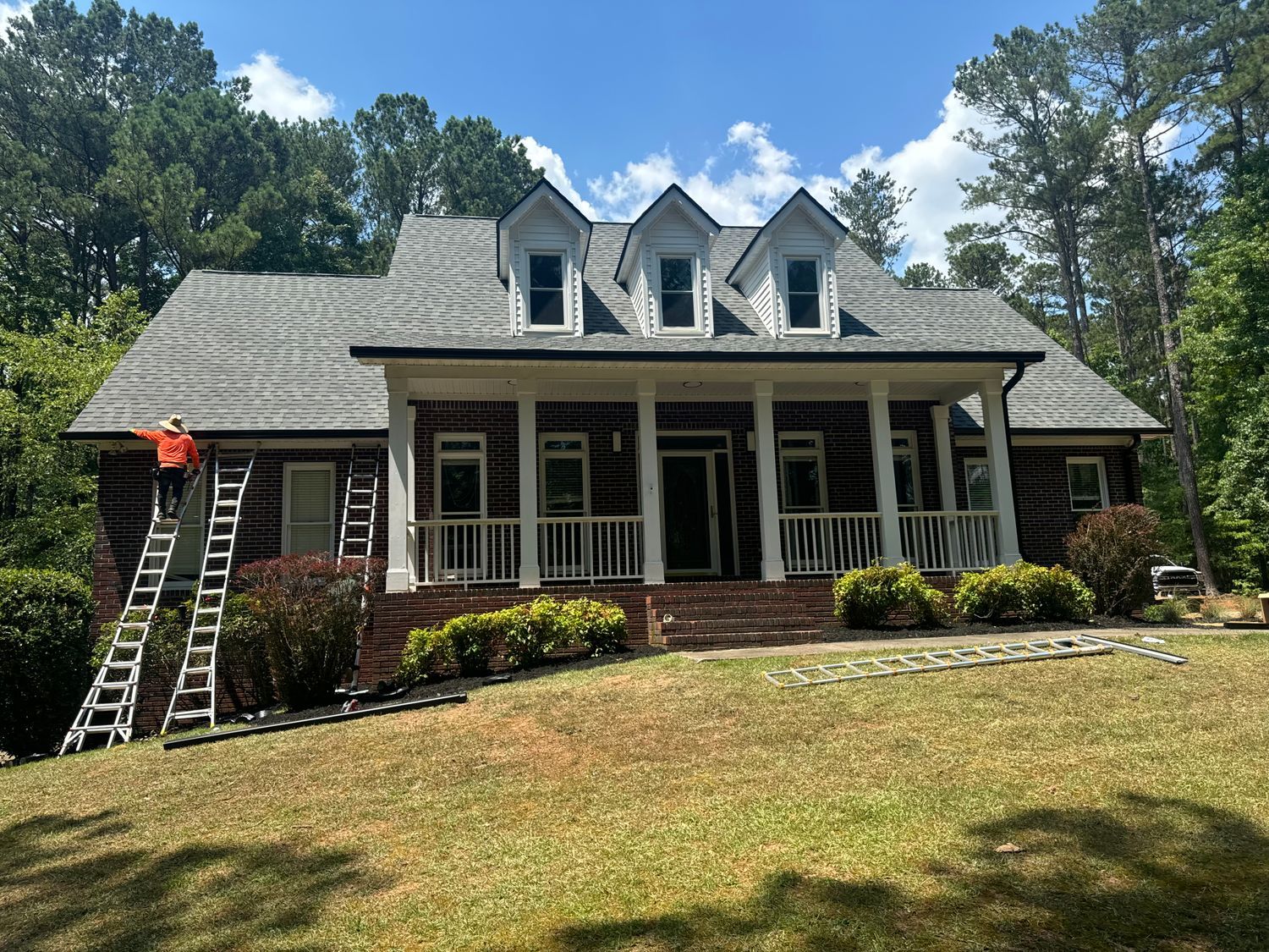 Person on a ladder near a brick house with white trim and a covered porch on a sunny day.