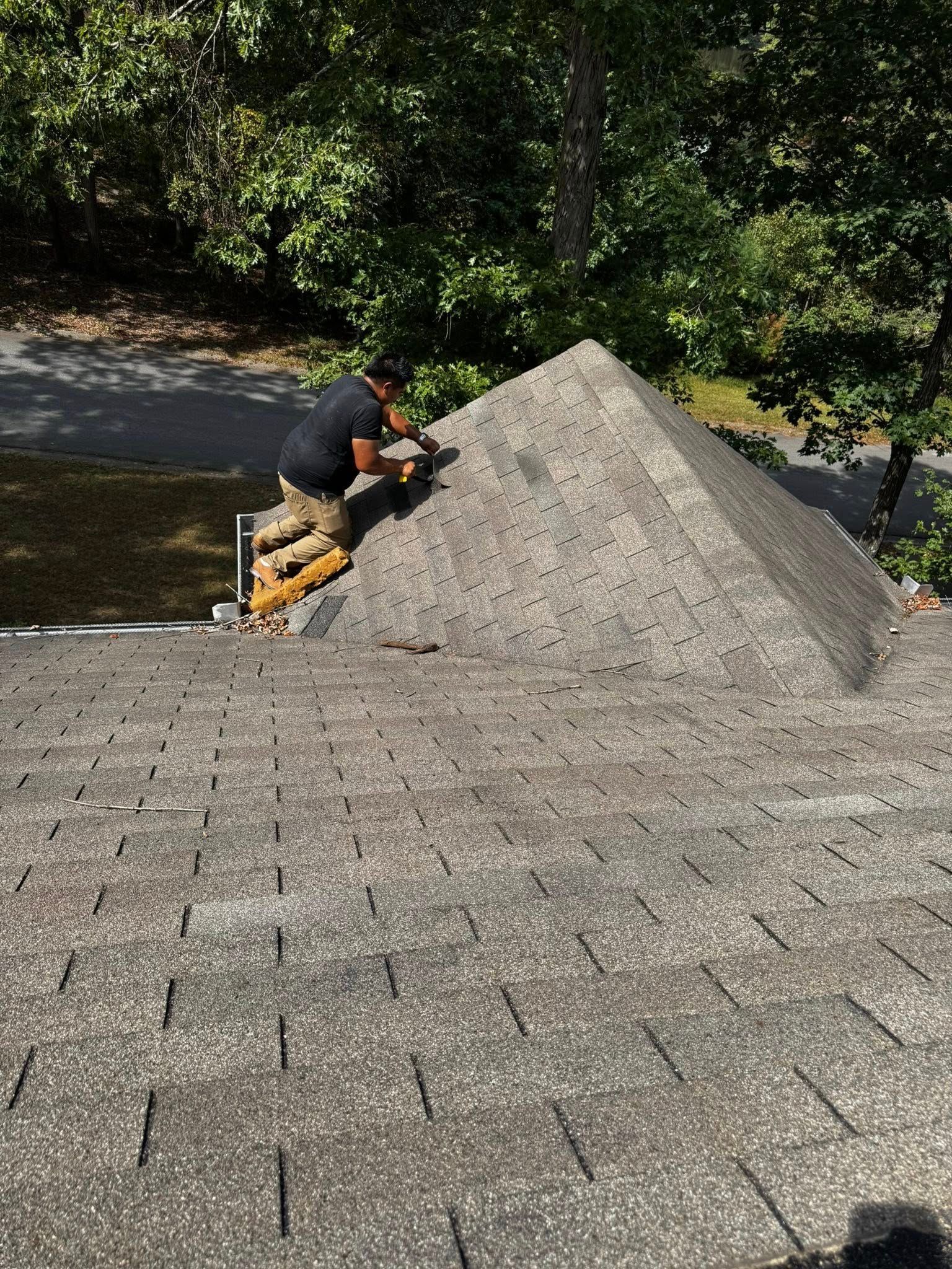 Roofer working on a gray shingled roof, trees in the background, sunny day.