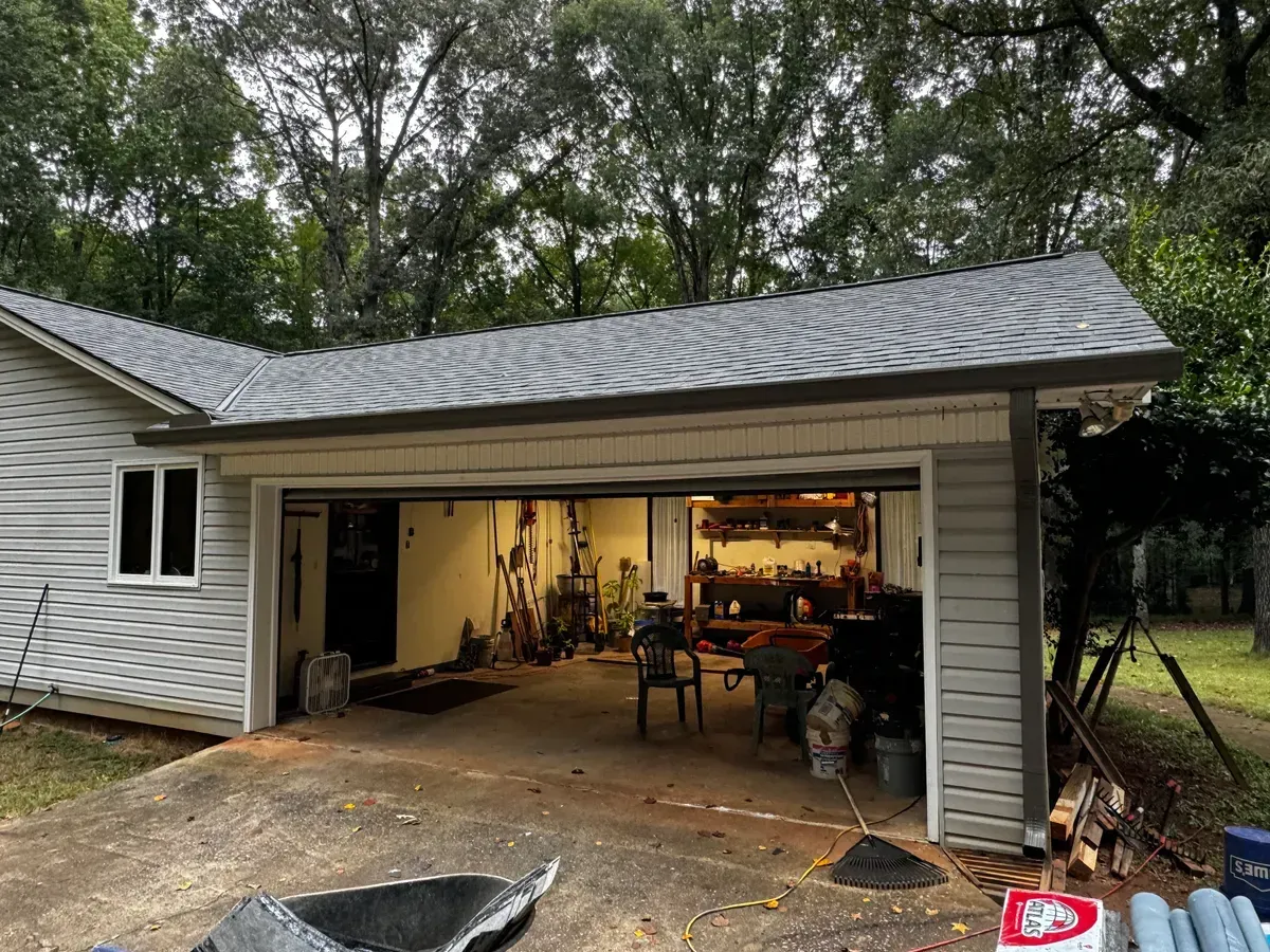 Garage with open door, light gray siding, dark gray roof, and cluttered interior.