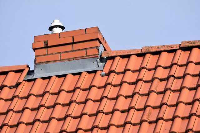 Red brick chimney on an orange tile roof, with a white vent cap, against a blue sky.