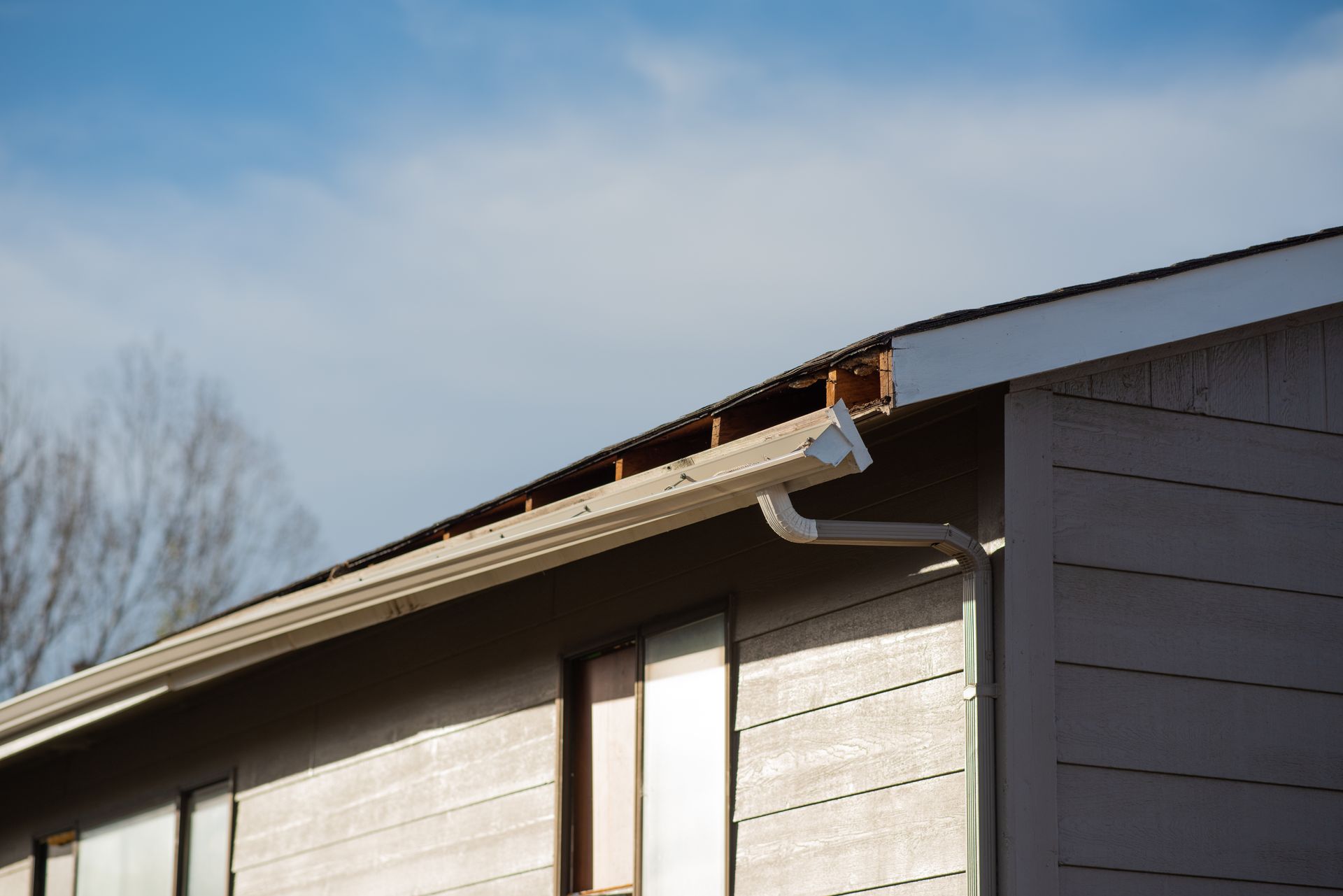 Damaged roof edge with missing wood and gutter; cloudy blue sky in background.