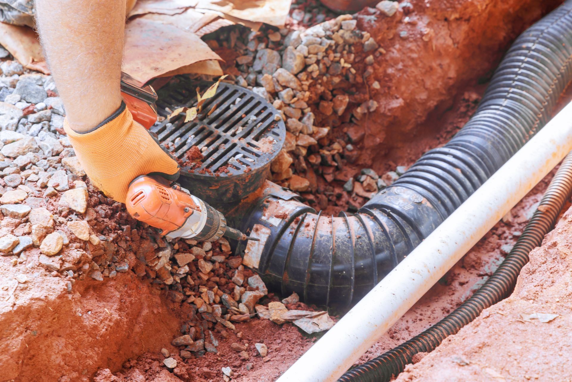 Person with orange gloves uses a drill to connect a drainpipe to a catch basin in a trench.