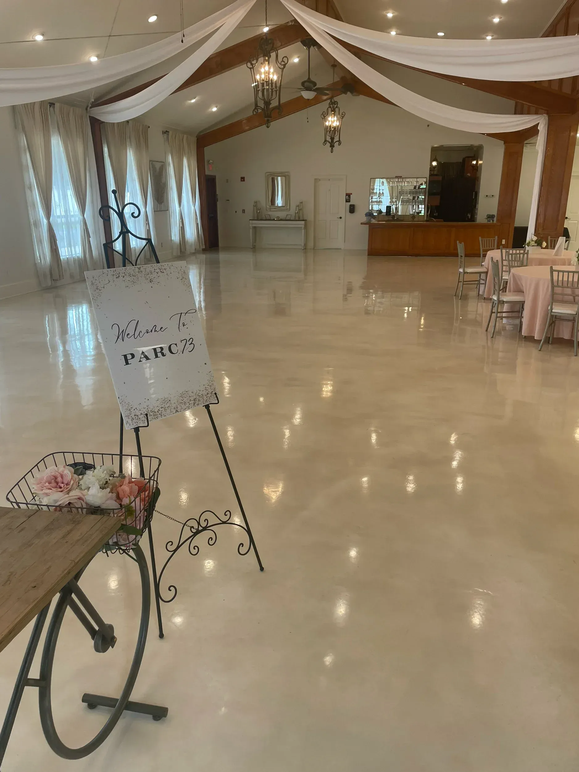 Empty event hall with light-colored polished floor, draped ceiling, and tables set. A welcome sign and decorative bicycle are in the foreground.