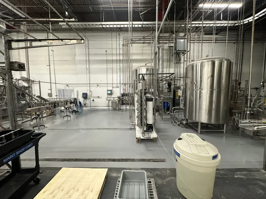 A factory interior with industrial equipment, tanks, and pipes. Gray floors, white walls.