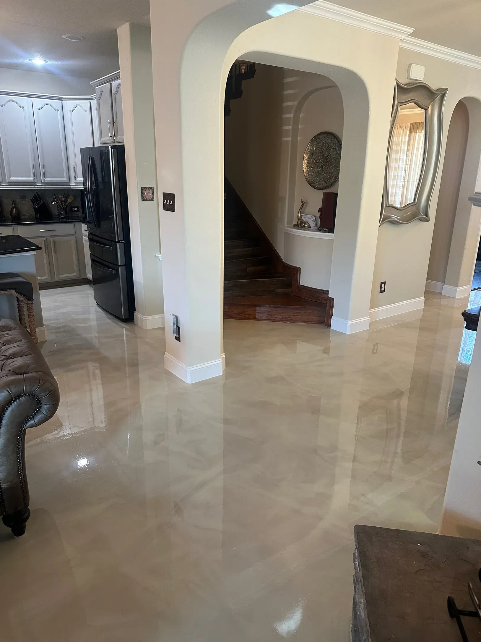 Shiny, light-colored epoxy flooring in a home. Arches lead to a staircase and kitchen with black appliances.