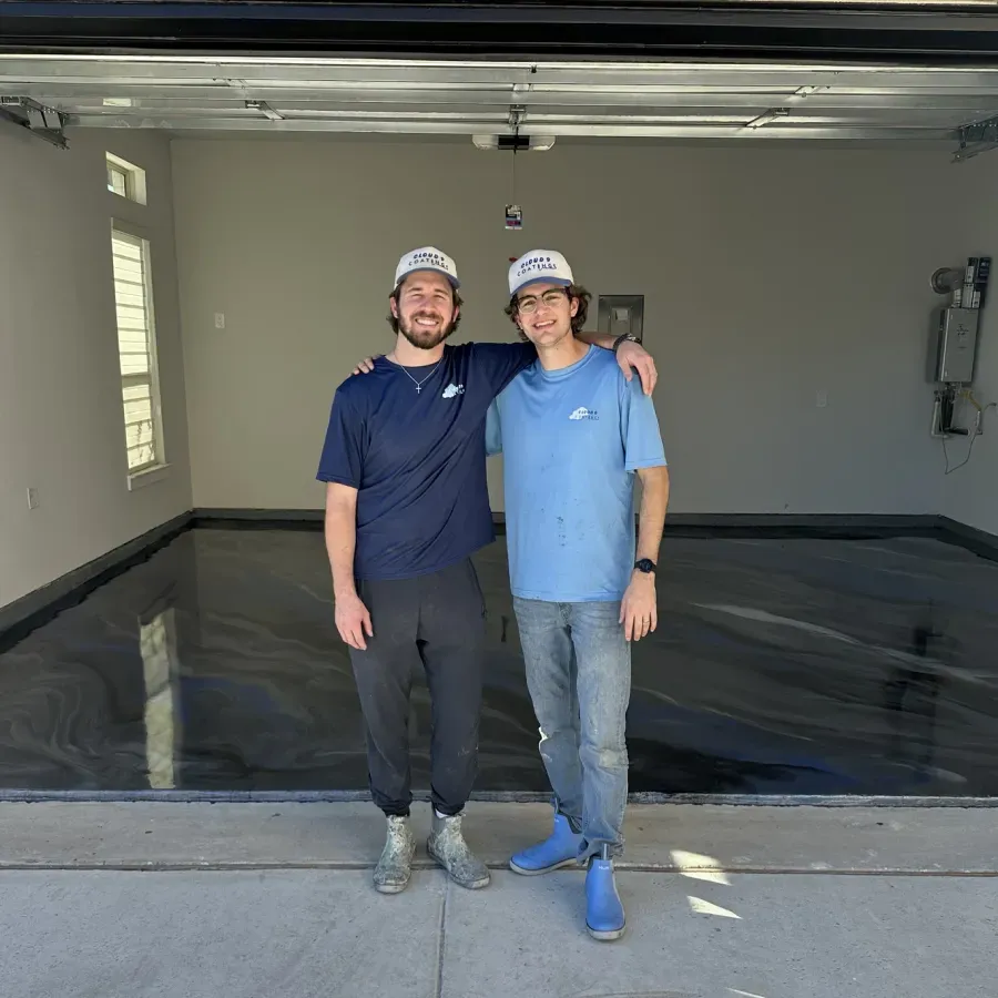 Two men stand in a finished garage with a metallic epoxy floor. Both wear hats and t-shirts, posing with arms around each other.
