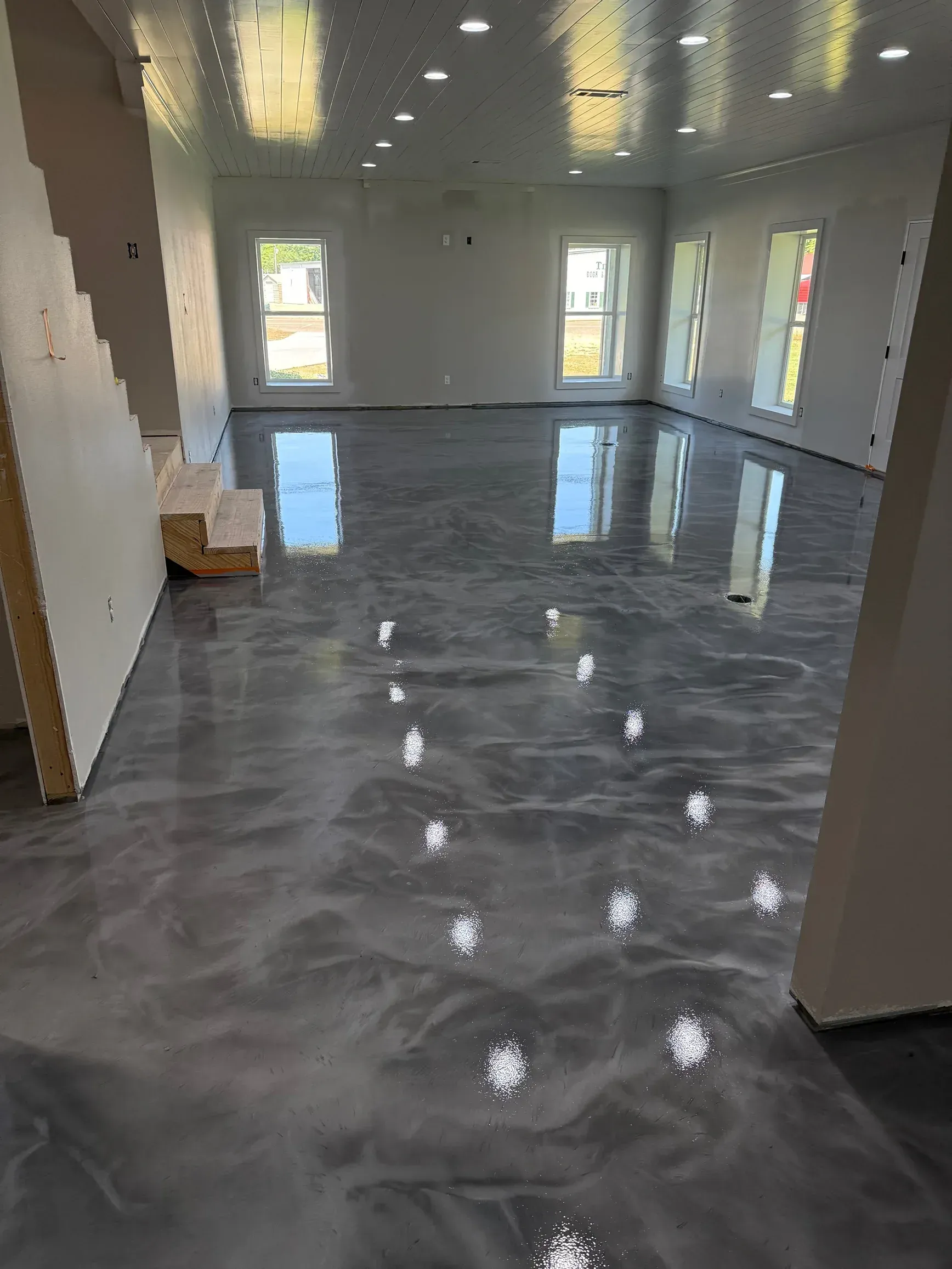 Spacious room with shiny, gray epoxy floor. Windows reflect light. Stairs visible on the left side.