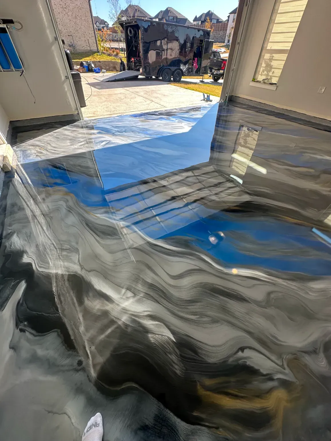Epoxy-coated garage floor with black, gray, and blue swirling patterns reflecting sunlight, with a construction site in the background.