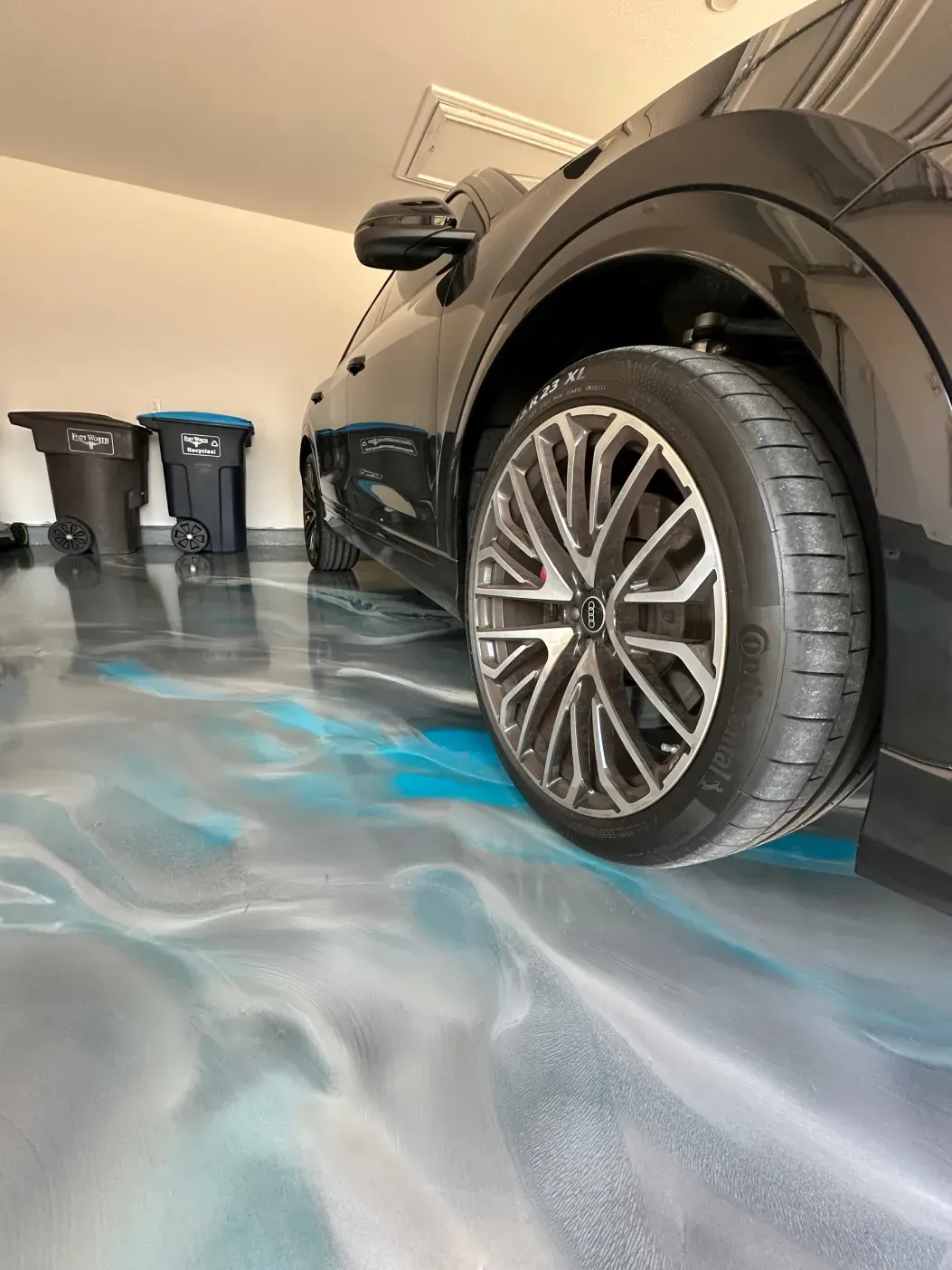 Black car parked in garage with blue and silver epoxy floor; trash cans in background.