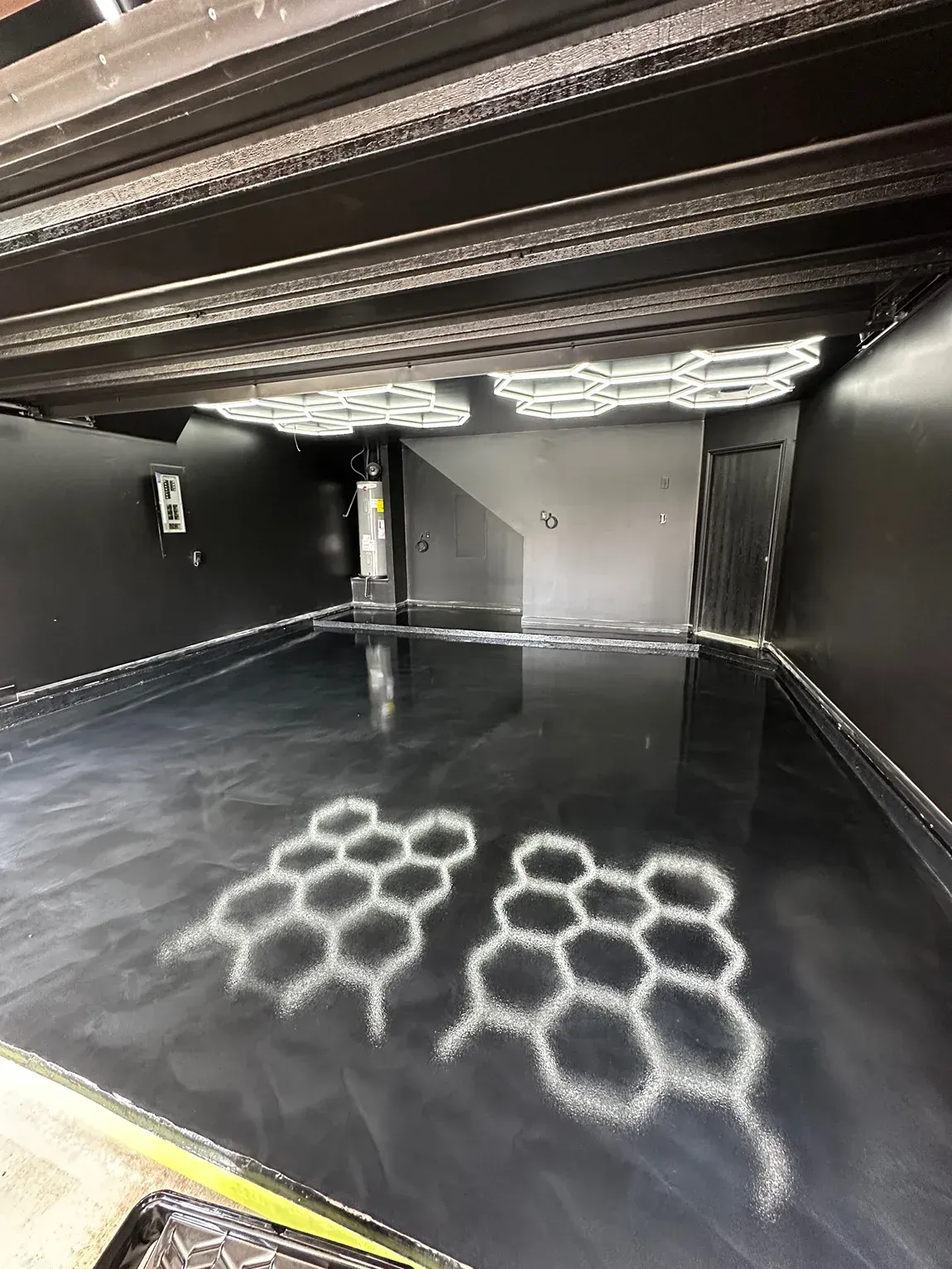 Black walled room with reflective floor, decorated with white honeycomb shapes; lights on ceiling.
