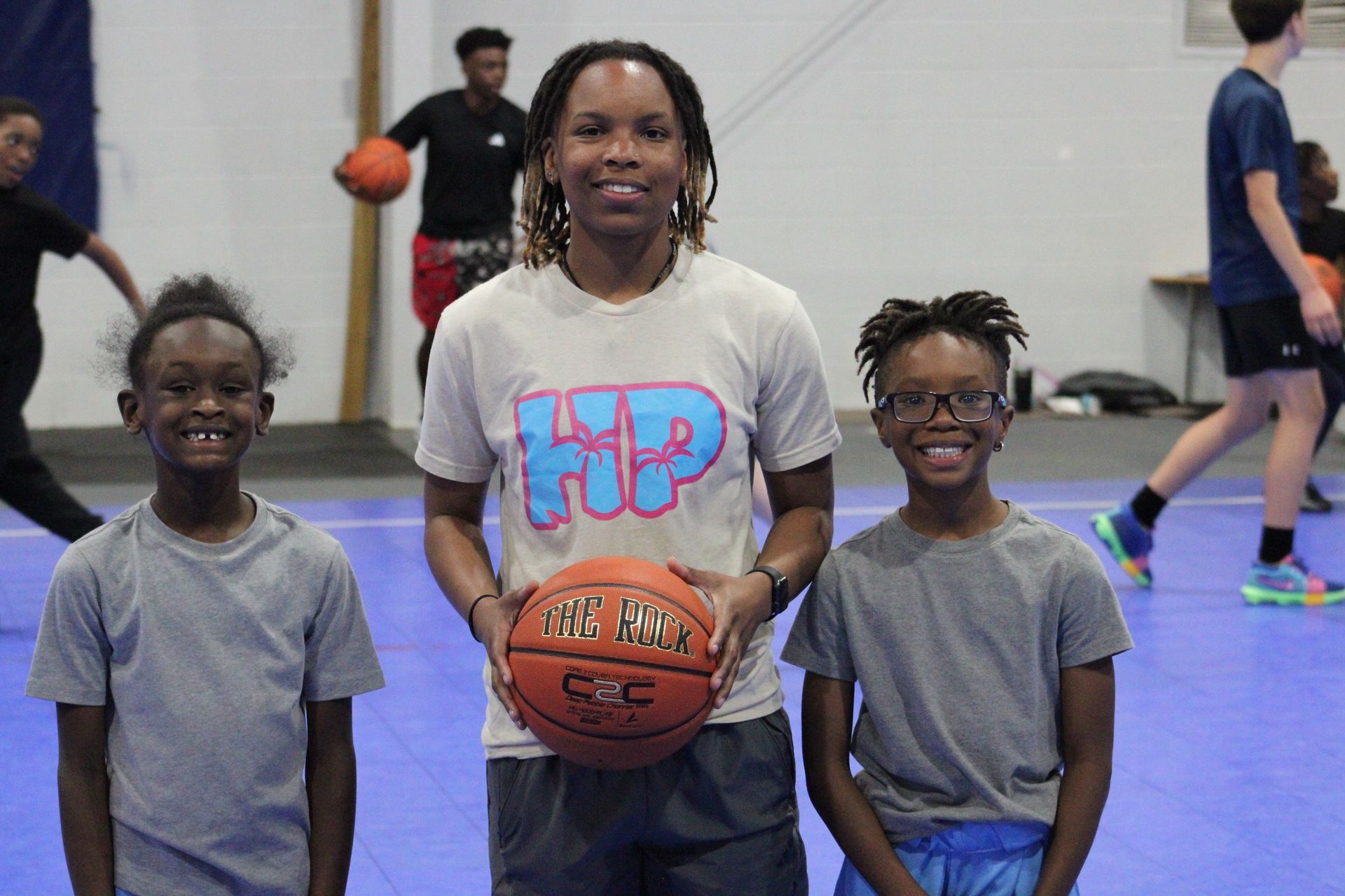 A person stands holding a basketball between two others on a blue indoor court, all smiling at the camera.