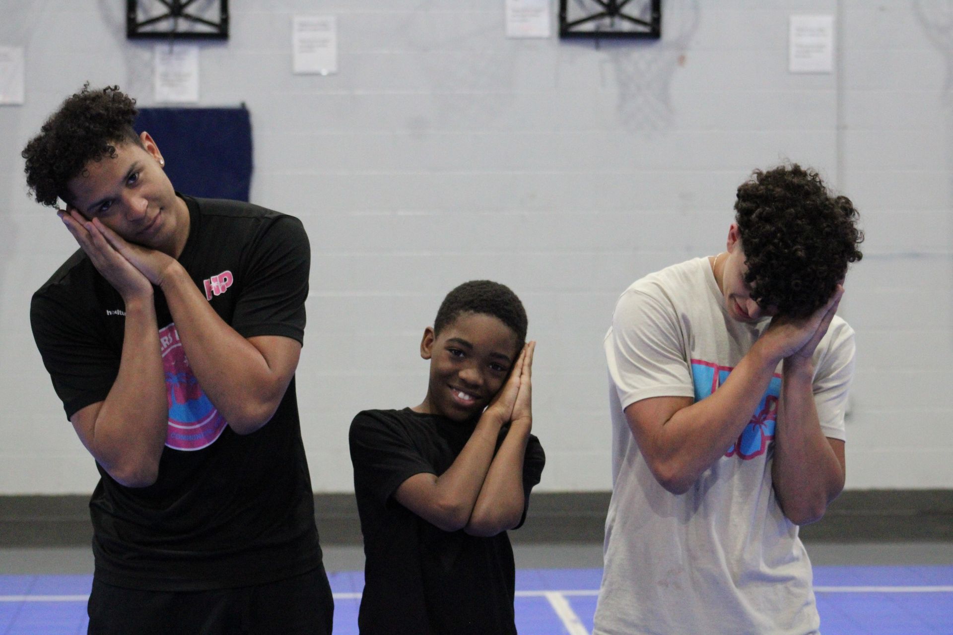 Three people in an indoor gym pose with their hands pressed against their cheeks as if sleeping.