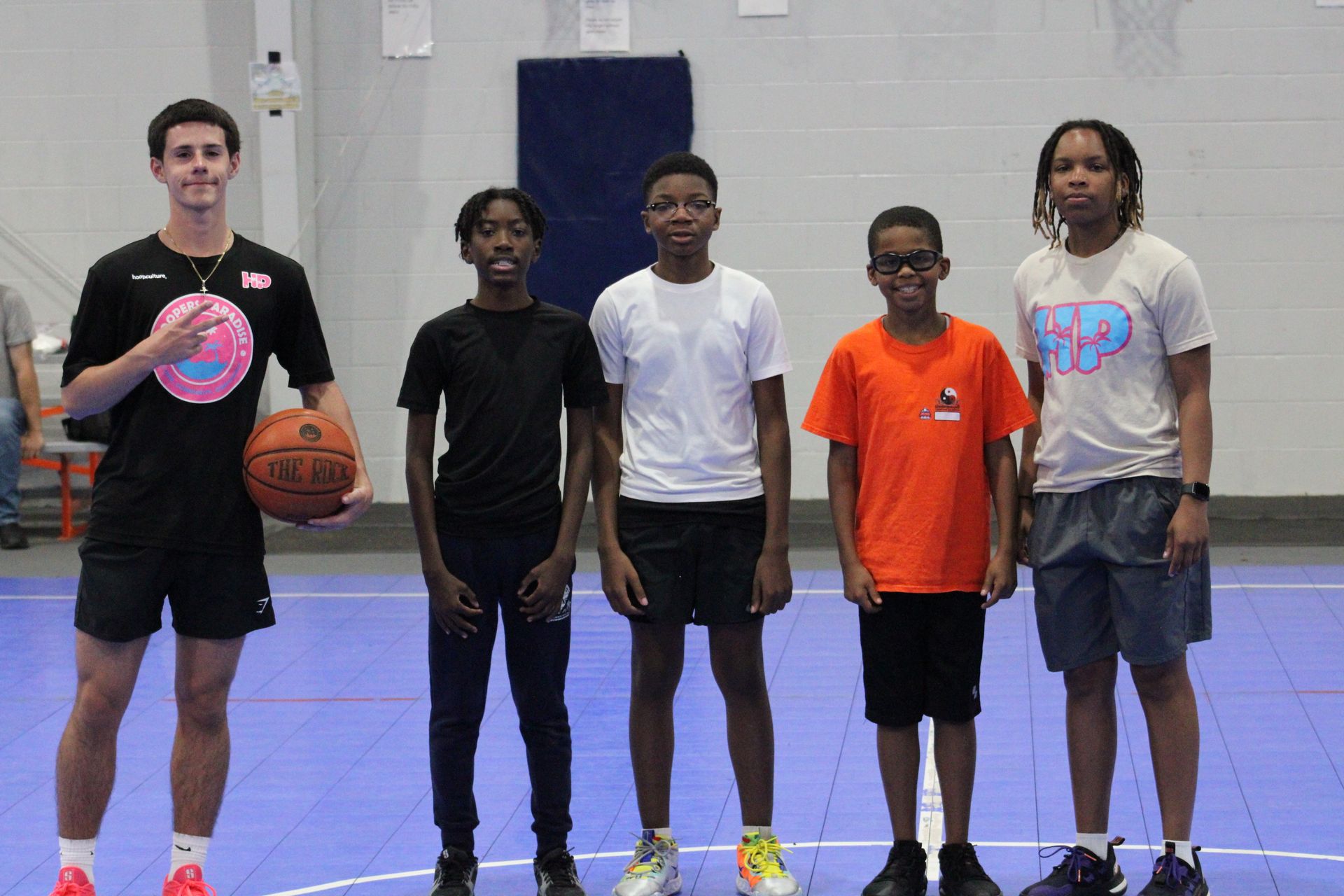 Five people stand in a line on a blue basketball court, one holding a basketball, against a white wall.